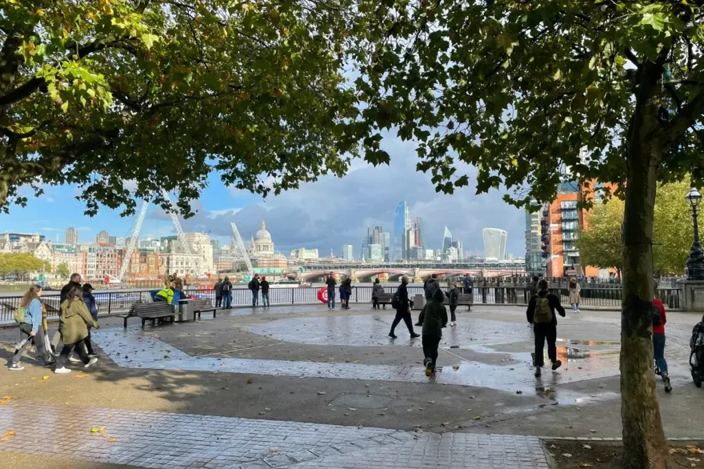 People milling around on the South Bank around the River Thames with the London skyline including St Paul's and The Shard behind them.