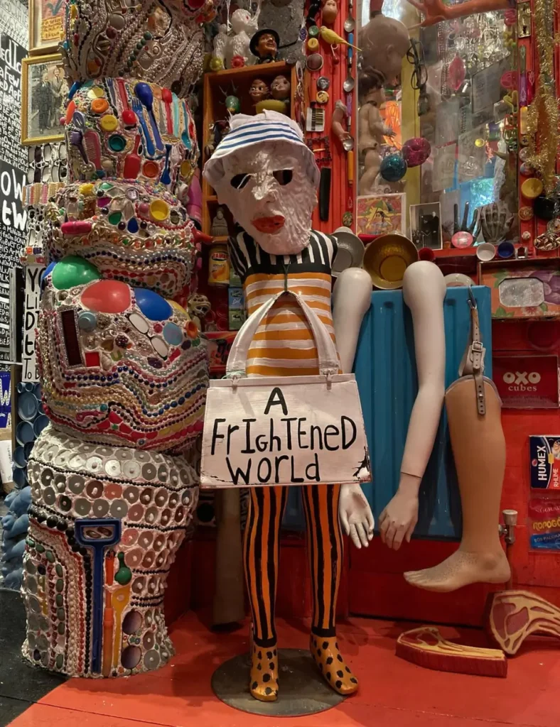 Masked mannequin holding an โA Frightened Worldโ sign next to a mosaic totem at House of Dreams Museum, East Dulwich